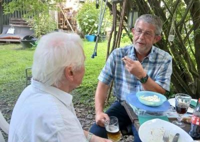 Zwei ältere Herren sitzen an einem Tisch im Garten und genießen gemeinsam eine Mahlzeit bei sonnigem Wetter.