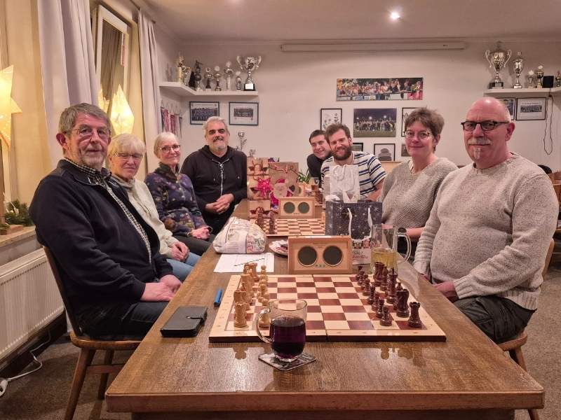 Acht Mitglieder des Schachclub Büsum beim Jul-Klapp-Turnier 2025. Vier Personen sitzen auf jeder Seite eines Tisches, vor ihnen liegen Schachbretter, Geschenke und Pokale sind im Raum sichtbar.